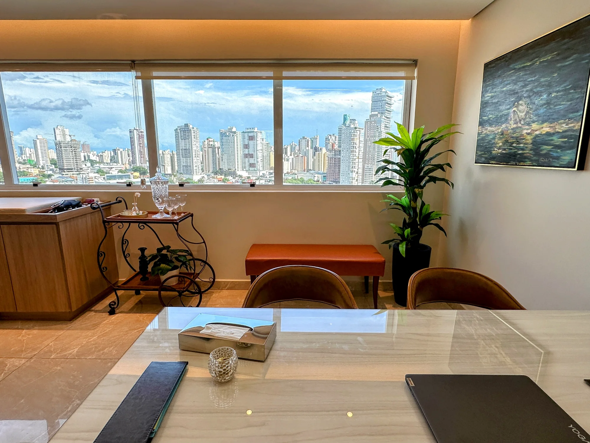 A mesa de pedra clara em primeiro plano e, ao fundo, a janela larga com o horizonte de prédios de Goiânia; à esquerda, o carrinho de ferro com cristais; ao centro, um banco terracota e uma planta; na parede, um quadro.