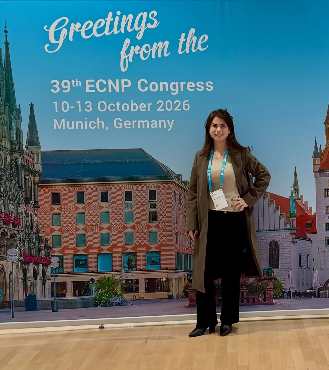 Dra. Daniela Londe de pé, com o crachá do congresso, diante do painel “Greetings from the 39th ECNP Congress, 10-13 October 2026, Munich, Germany”.