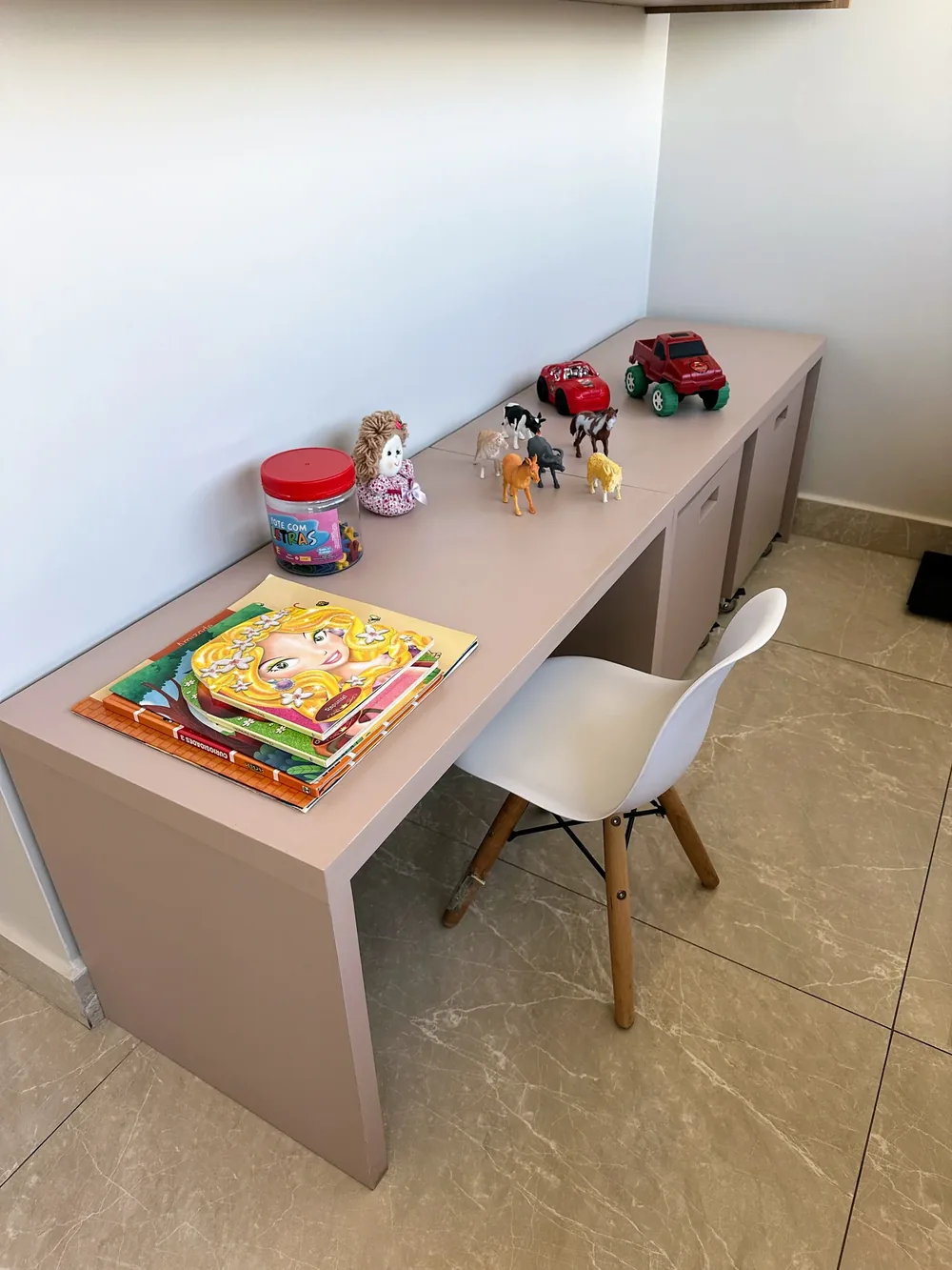 Bancada baixa com livros infantis, bonecos, bichinhos e carrinhos de brinquedo, e uma cadeira pequena branca.