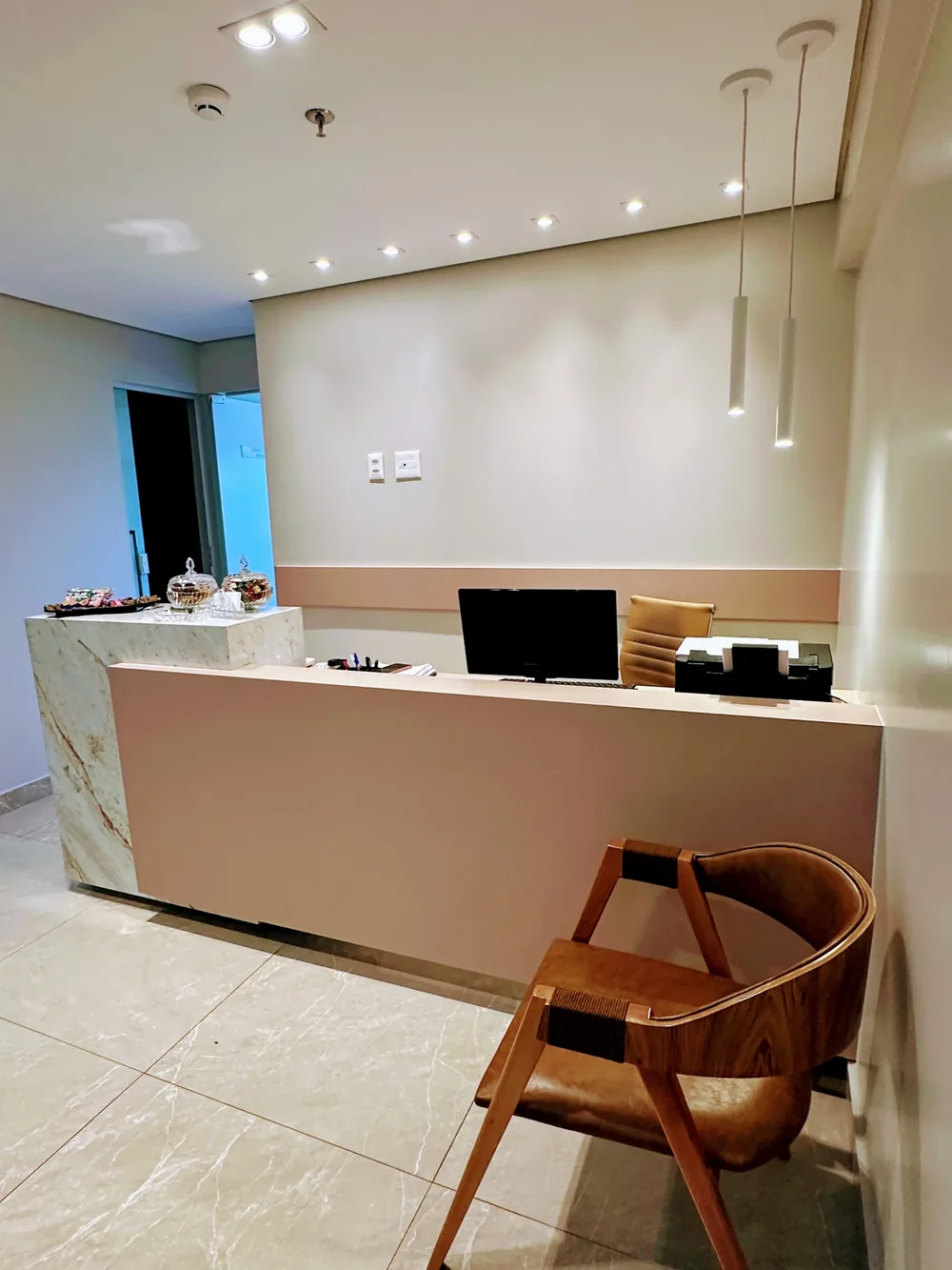 A recepção: balcão de pedra clara com potes de cristal com balas, luminárias pendentes e uma cadeira de madeira e couro.