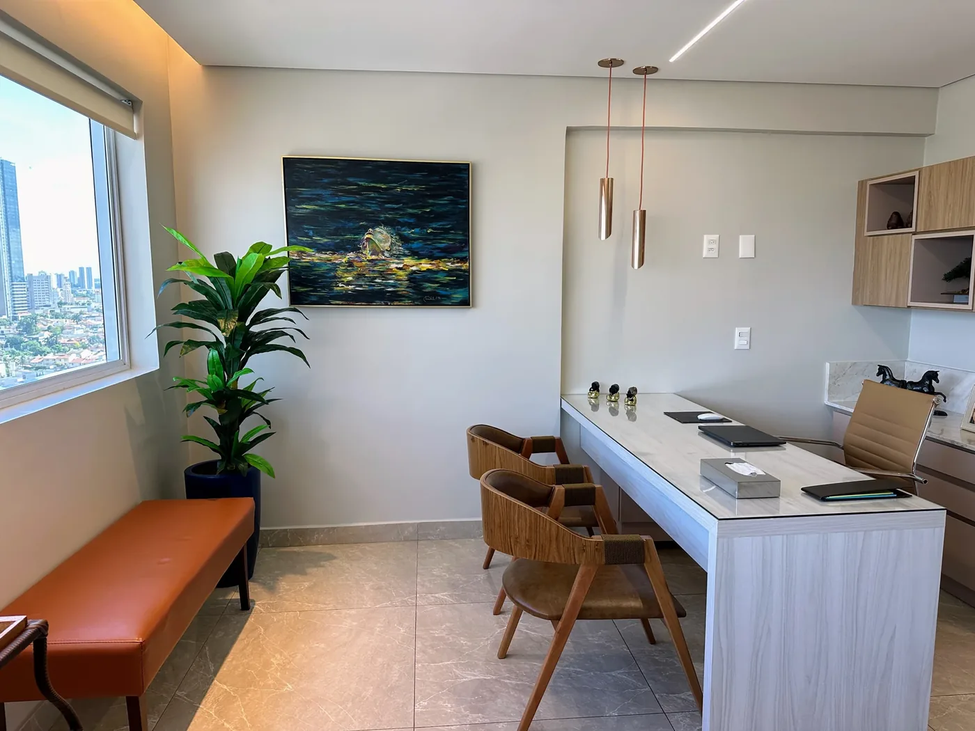 A sala vista de lado: um quadro de tons escuros na parede, a mesa de pedra com duas cadeiras de madeira e couro, uma planta e o banco terracota sob a janela.